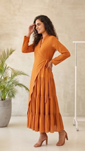 Woman wearing an orange dress standing in a room with a plant and a coat rack.